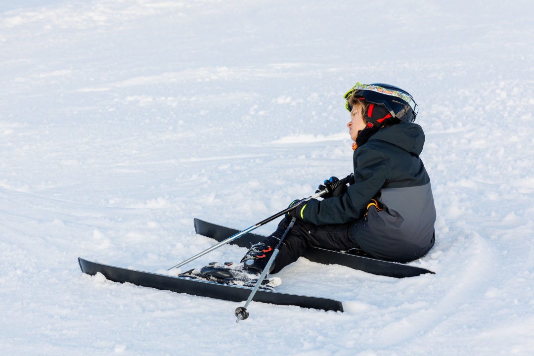 Little teen boy kid in black suit lying on snow after ski downhill accident crash. Tired beginner child learn skiing at winter ski camp school on mountain resort.