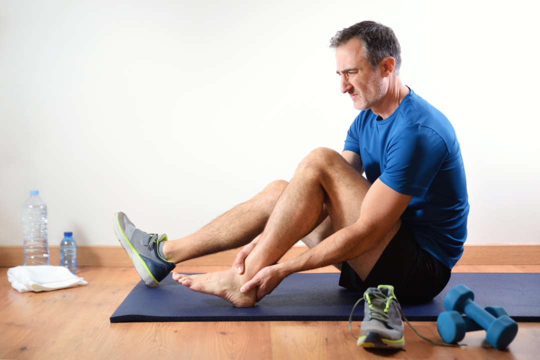 Man sitting on a mat complaining of ankle pain after doing indoor sports exercises. Side view.