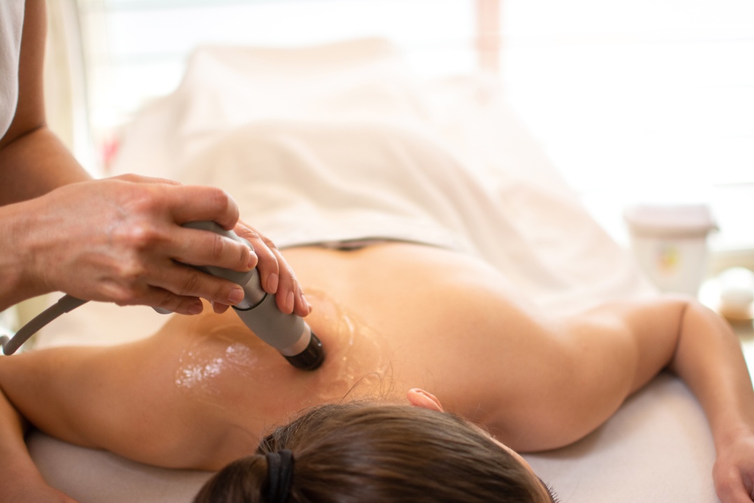 Close up of extracorporeal shockwave therapy in beauty salon. Physical therapy for neck and back muscles,spine with shock waves. Young woman wearing protective mask during treatment in salon.